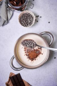 A metal spoon sprinkling chocolate chips into a saucepan filled with milk and cocoa powder. Part of a piece of baking chocolate and a jar of chopped chocolate can be seen on the sides. A small bowl of chocolate chips is next to the saucepan and a few chocolate chips are sprinkled throughout the table.