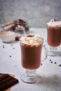 A glass mug of hot chocolate decorated with whipped cream and chocolate shavings. Another glass mug of hot chocolate can be seen behind it. In the foreground is a piece of baking chocolate and chocolate chips are scattered on the table. In the background are small container of chopped chocolate, chocolate chips, and maple syrup.