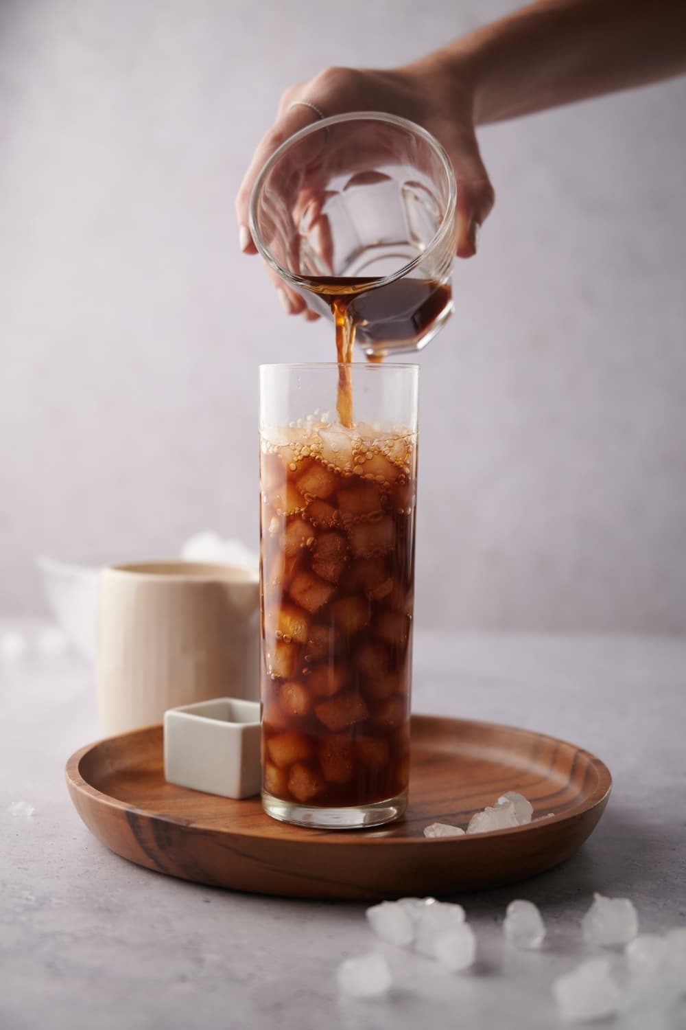 A hand pouring coffee from a small glass into a taller glass of ice. The glass is on a wooden board decorated with scattered pieces of ice. A small square bowl is next to the glass and a ceramic milk pot is behind it.