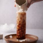 A hand pouring almond milk from a small ceramic milk pot into a tall glass of ice and coffee. The glass is on a wooden tray decorated with scattered piece of ice and a bowl with more ice can be seen in the back.