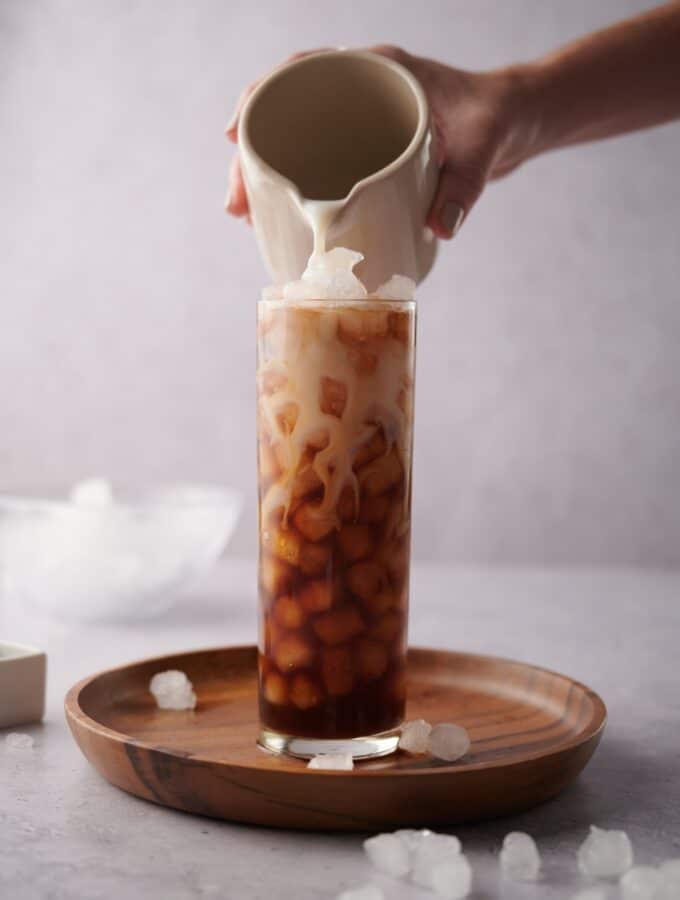 A hand pouring almond milk from a small ceramic milk pot into a tall glass of ice and coffee. The glass is on a wooden tray decorated with scattered piece of ice and a bowl with more ice can be seen in the back.