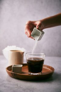 A hand pouring granulated sweetener from a small square container into a small glass of coffee. The glass is on a wooden board next to another small square container holding vanilla extract. A small milk pot and a bowl of ice are in the back.