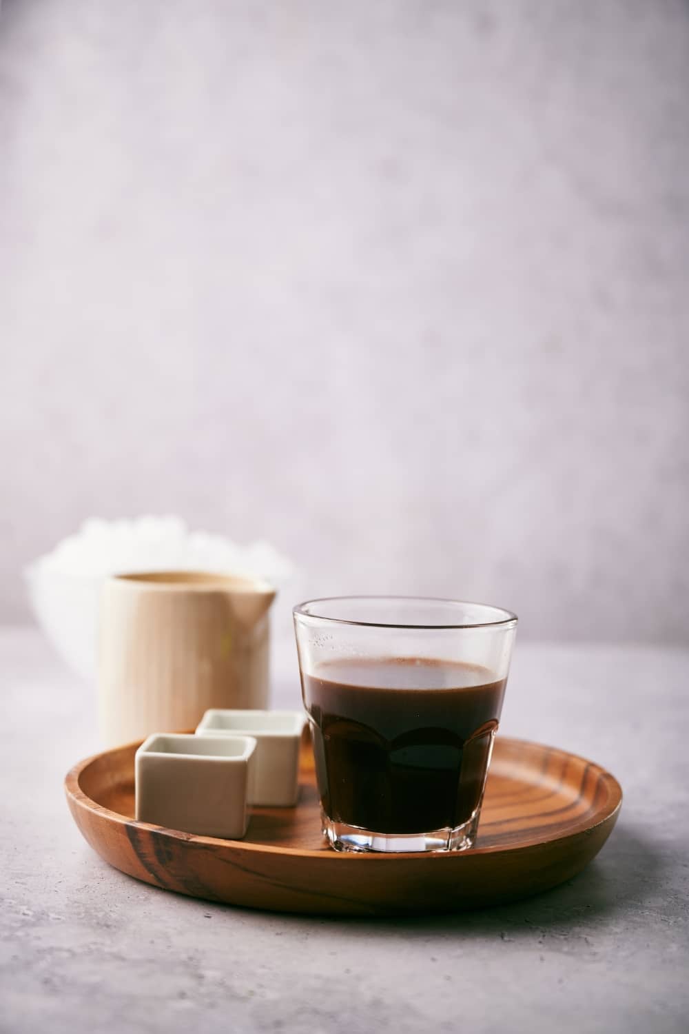 A small glass of coffee next to two small square containers on a wooden tray. A small milk pot is in the back in front of a bowl of ice.