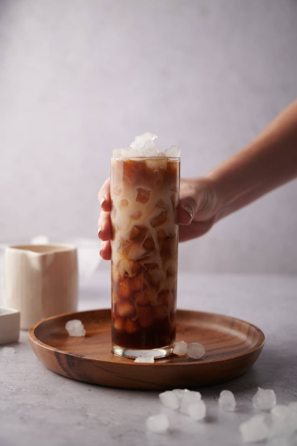 A hand holding a tall glass of iced vanilla latte on a wooden tray. The milk and coffee are swirled in the glass and the tray is decorated with scattered ice. A small milk pot sits in the back.