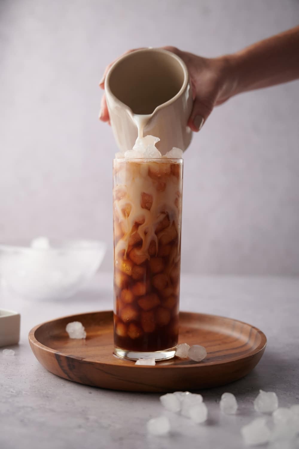 A hand pouring almond milk from a small ceramic milk pot into a tall glass of ice and coffee. The glass is on a wooden tray decorated with scattered piece of ice and a bowl with more ice can be seen in the back.