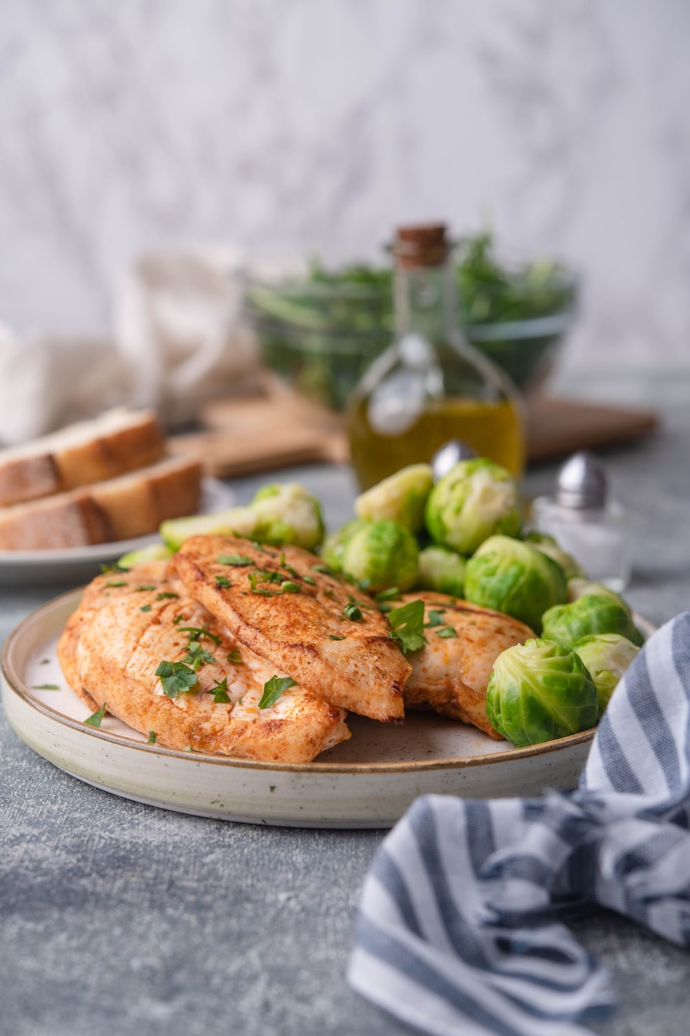 Sauteed chicken breasts garnished with herbs on a plate with steamed brussel sprouts. Surrounding the plate is a striped tea towel, a plate of sliced bread, a bottle of olive oil, salt and pepper shakers, and a glass bowl of greens next to a beige tea towel.