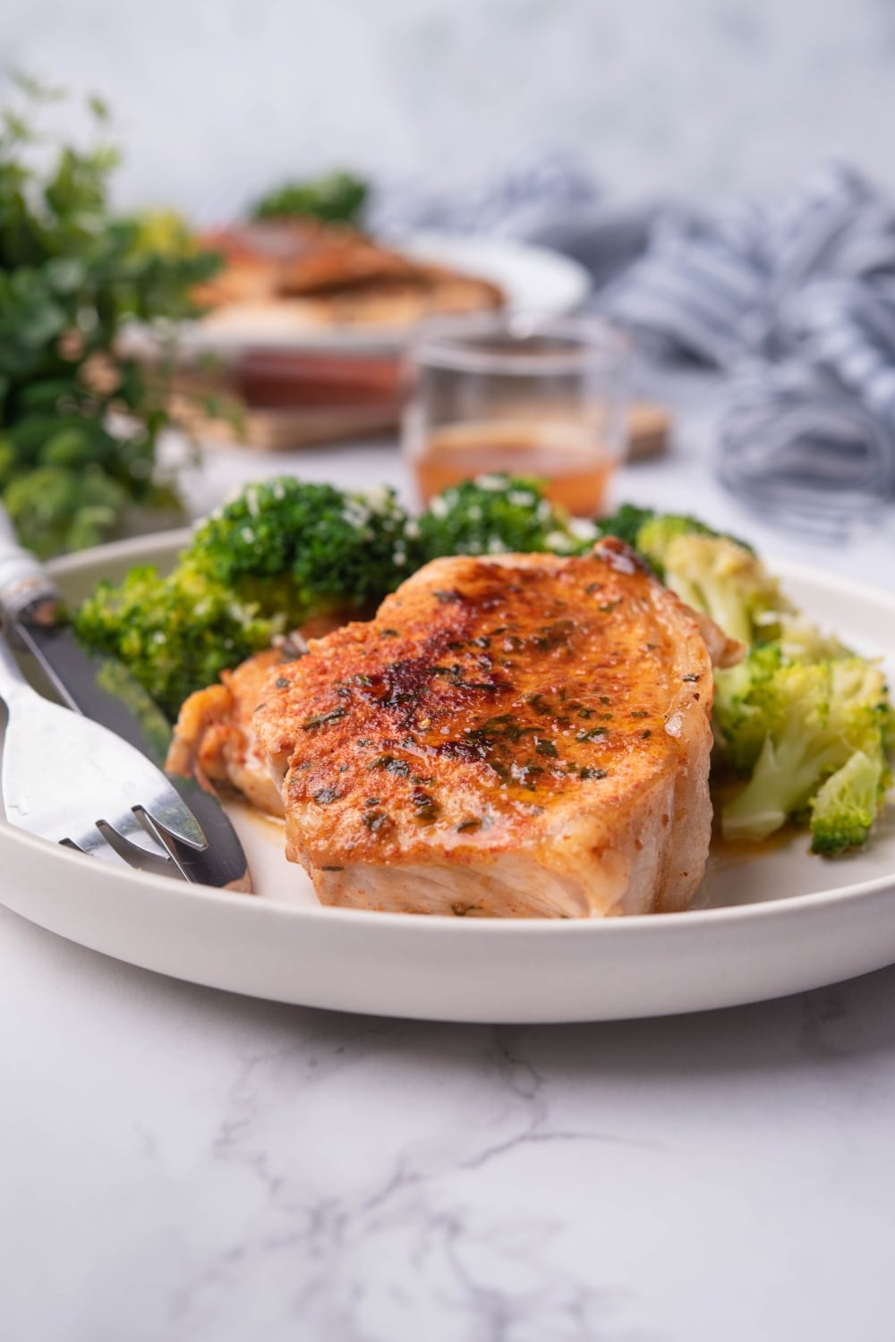 Pan seared pork chops with broccoli on a white plate. A white handled fork and knife are on the plate and another plate of pork chops and broccoli can be seen in the background next to a glass sauce pitcher of pan sauce.