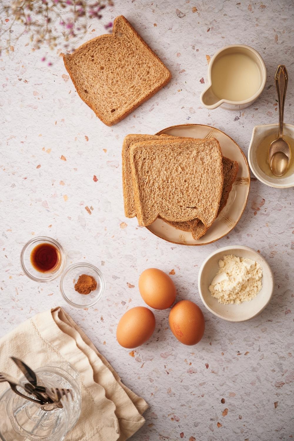 Clockwise from top: a slice of wheat bread, a small pitcher of almond milk, a small pitcher of syrup with a metal spoon, a plate with two wheat bread slices, a small bowl of protein powder, three eggs, and two small glass bowls of cinnamon and vanilla extract.