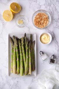 A plate of raw asparagus on a white rectangle plate, a halved lemon, a small bowl of garlic paste, a medium bowl of panko breadcrumbs, a small bowl of butter, and salt and pepper shakers.