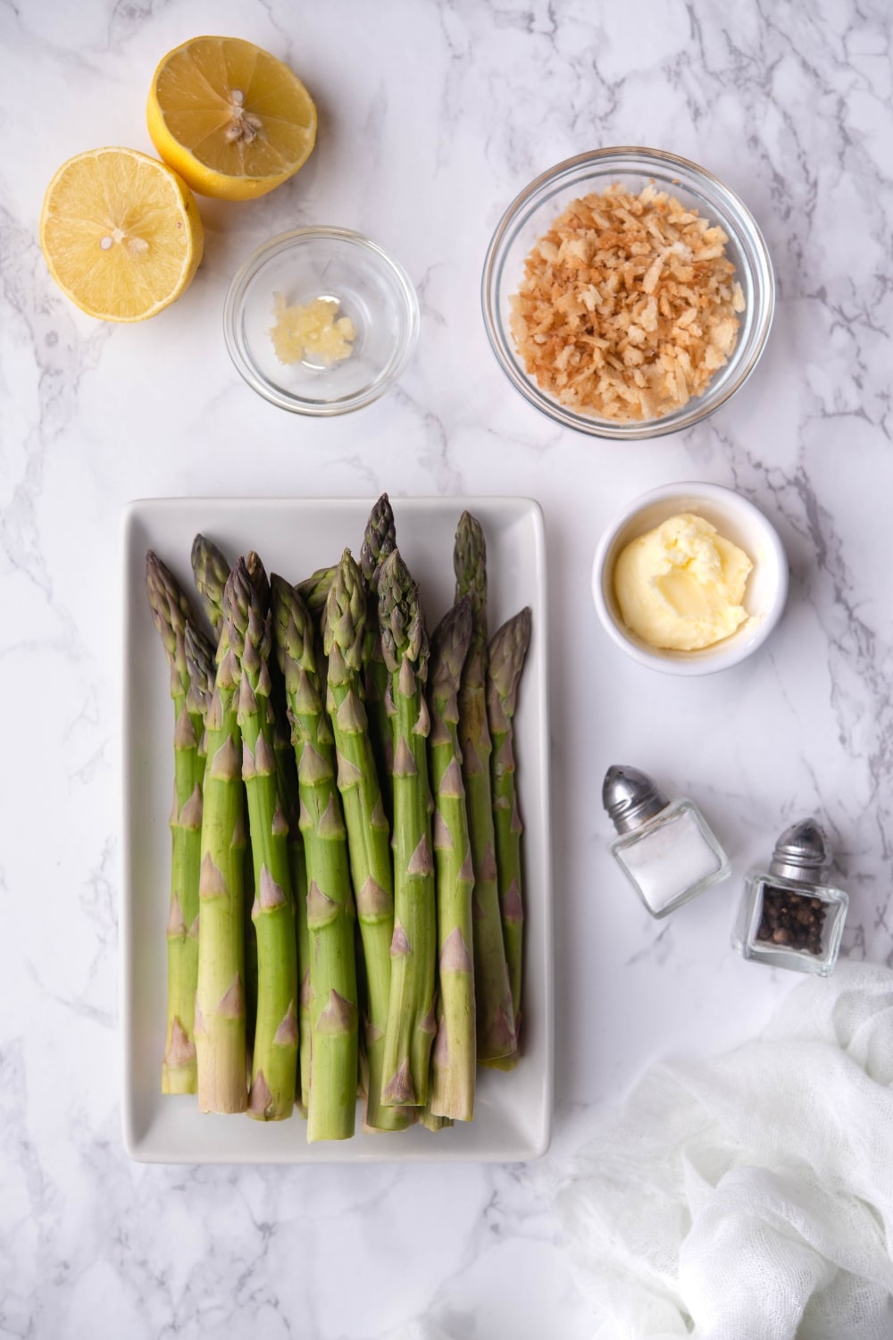 A plate of raw asparagus on a white rectangle plate, a halved lemon, a small bowl of garlic paste, a medium bowl of panko breadcrumbs, a small bowl of butter, and salt and pepper shakers.