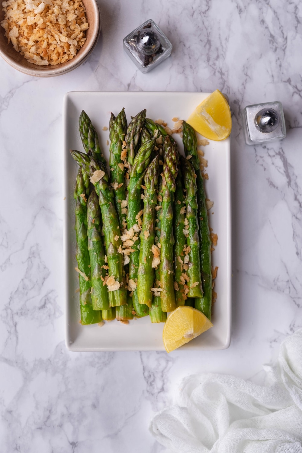 Garlic butter sauteed asparagus topped with panko breadcrumbs and served with lemon wedges on a white rectangle plate. Salt and pepper shakers and part of a small bowl of breadcrumbs surround the plate.