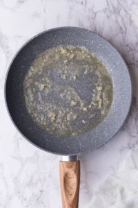 Garlic paste sizzling in a pan of melted butter. The pan is speckled grey with a wooden handle on a marble countertop.