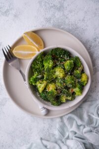 Top view of sauteed broccoli florets in a white bowl on top of an ecru colored plate. Next to the bowl, there are two lemon wedges and a fork with a white handle.