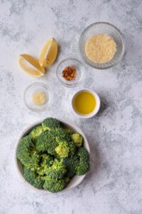 Sauteed broccoli ingredients. Two lemon wedges, a glass bowl of grated parmesan, a white bowl of broccoli florets, small glass bowls 0f garlic paste and red pepper flakes, and a white ramekin with olive oil.