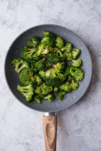 Sauteed broccoli in a grey speckled skillet with a wooden handle. A small amount of garlic paste is in the center of the skillet and red pepper flakes are sprinkled throughout.
