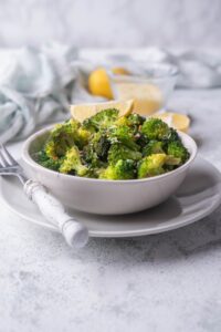 Sauteed broccoli florets with red pepper flakes and grated parmesan in a white bowl garnished with a lemon wedge. The bowl is on a white plate next to a fork with a white handle. Behind it are more lemon wedges and a gently crumpled tea towel.