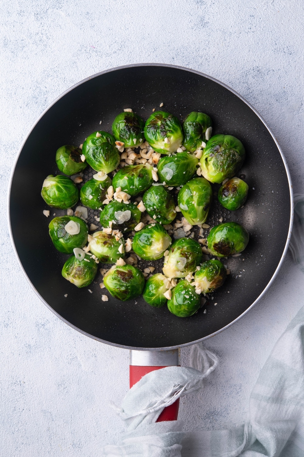 Sauteed brussels sprouts tossed with garlic slices and chopped hazelnuts in a black pan. A tea towel is wrapped around the handle of the pan.