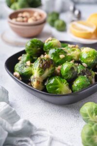 A closer look at sauteed brussels sprouts garnished with hazelntus in a small square cast iron skillet. Behind them is a bowl of hazelnuts, salt and pepper shakers, a plate of lemon with half a lemon wedge, and part of a bowl of fresh raw brussels sprouts.
