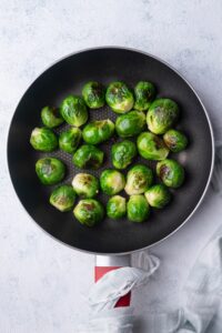 Sauteed brussels sprouts in a black pan. A tea towel is wrapped around the handle of the pan, which is resting on a grey countertop.