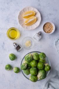 A large glass bowl of raw brussels sprouts, a small plate with two lemon wedges, small bowls of chopped blanched hazelnuts, olive oil, and garlic slices, and salt and pepper shakers.