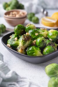 Close up of sauteed brussels sprouts in a small square cast iron skillet. The brussels sprouts are garnished with chopped hazelnuts. Behind them is a bowl of fresh raw brussel sprouts, a bowl of hazelnuts, salt and pepper shakers, and a plate of lemon with half a lemon wedge.