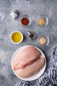 Sauteed chicken breast ingredients: salt in a salt shaker, three small glass bowls of paprika, garlic powder, and onion powder, whole black peppercorns in a pepper shaker, a small white ramekin of olive oil, and a plate with two raw chicken breasts.