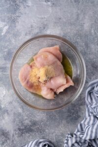 Raw chicken breasts slices in a glass bowl with olive oil and unmixed seasoning. The bowl is on a grey countertop next to a blue and white striped tea towel