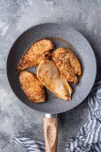 Four slices of sauteed chicken breasts in a grey speckled skillet with a wooden handle.