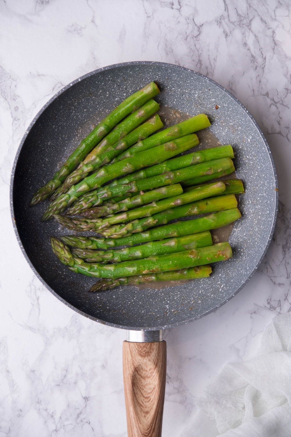 Sauteed asparagus with garlic and butter in a speckled grey pan with a wooden handle. The pan is on a marble countertop.