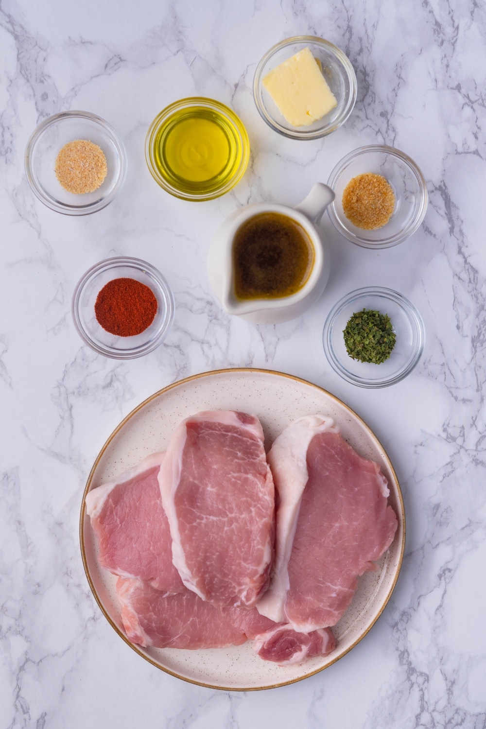 Raw pork chops on a white plate, a ceramic milk pitcher of chicken stock, and small glass bowls of butter, olive oil, garlic powder, onion powder, dried parsley, and paprika.