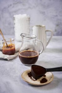 Skinny caramel macchiato ingredients. In the front is a plate holding a spoonful of coffee grounds. Close behind it is a small pitcher of cold brew coffee. Further back is a tall ceramic pitcher of milk, a small glass jar of caramel syrup, and a large mason jar filled with ice.