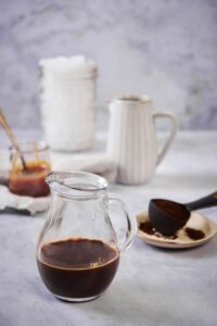 Skinny caramel macchiato ingredients. In the front is a pitcher of dark cold brew coffee with a plate holding a spoonful of coffee grounds behind it. Further back is a tall ceramic pitcher of milk, a small glass jar of caramel syrup, and a large mason jar filled with ice.