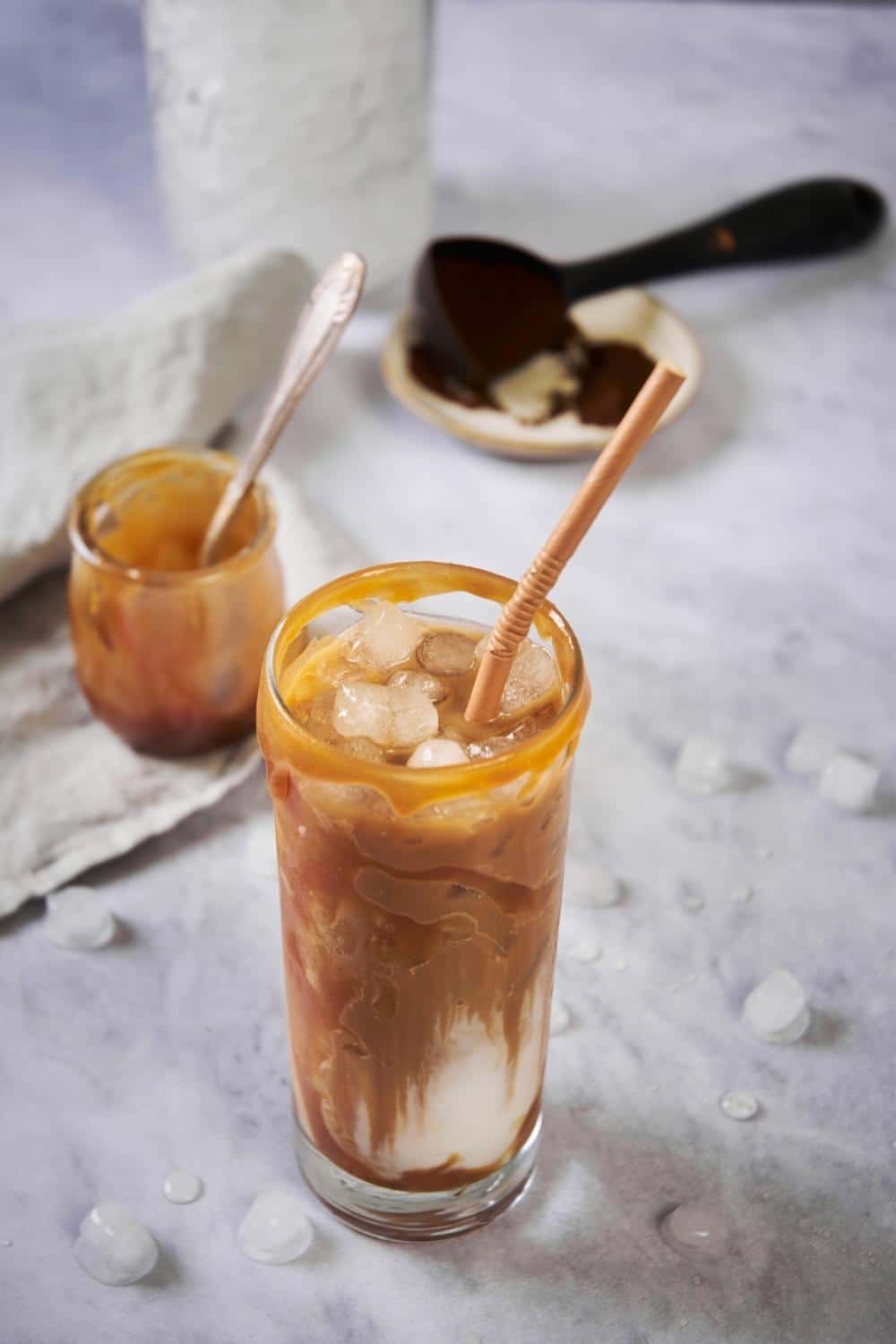 A tall glass of iced carame macchiato with a brown straw. Caramel syrup is drizzled on the rim and bits of ice can be seen floating in the swirled coffee and milk. Behind the glass is a small jar of caramel, a plate with a spoonful of coffee grounds, and a mason jar filled with ice.