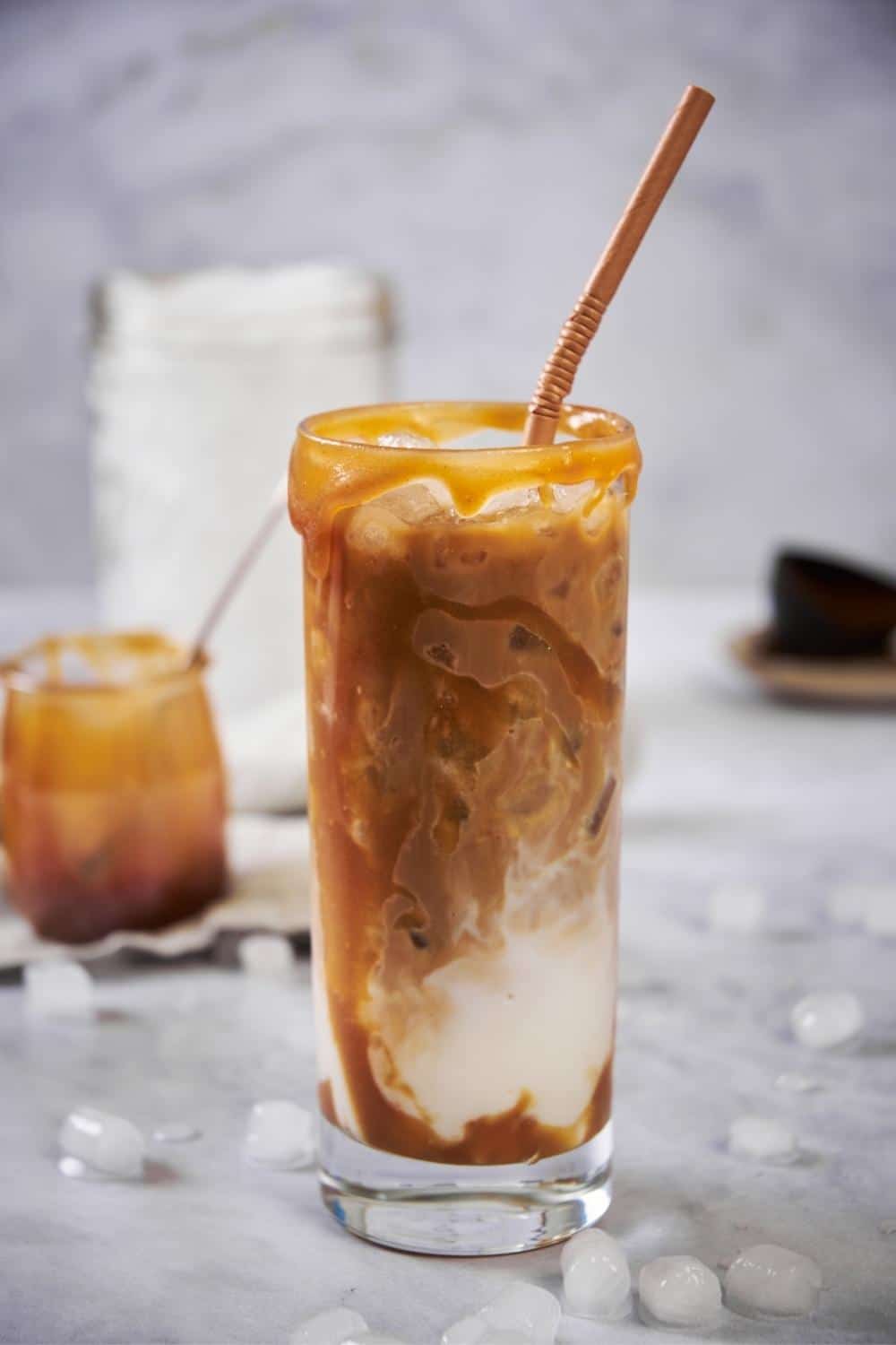 iced caramel macchiato in a tall glass with a brown straw. Caramel syrup is coating the rim and the insides of the glass and the milk and coffee are swirled. In the back is a large mason jar of ice and a small jar of caramel syrup.