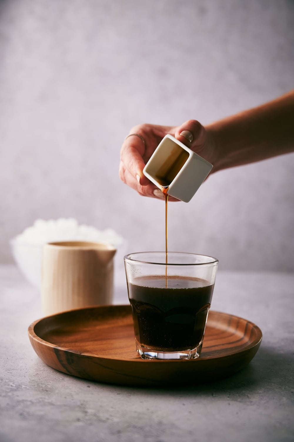 A hand pouring vanilla extract into a small glass of coffee. The glass is on a wooden tray and behind it is a small milk pot and a bowl of ice.