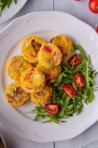 Two egg bites on top of six egg bites that are next to cut tomatoes on top of arugula on a white plate.