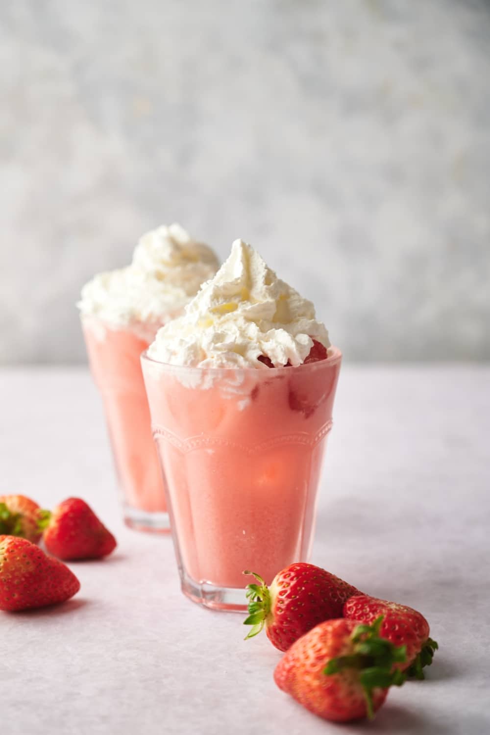 Two glasses filled with pink drink with whipped cream on top and some strawberries around them.
