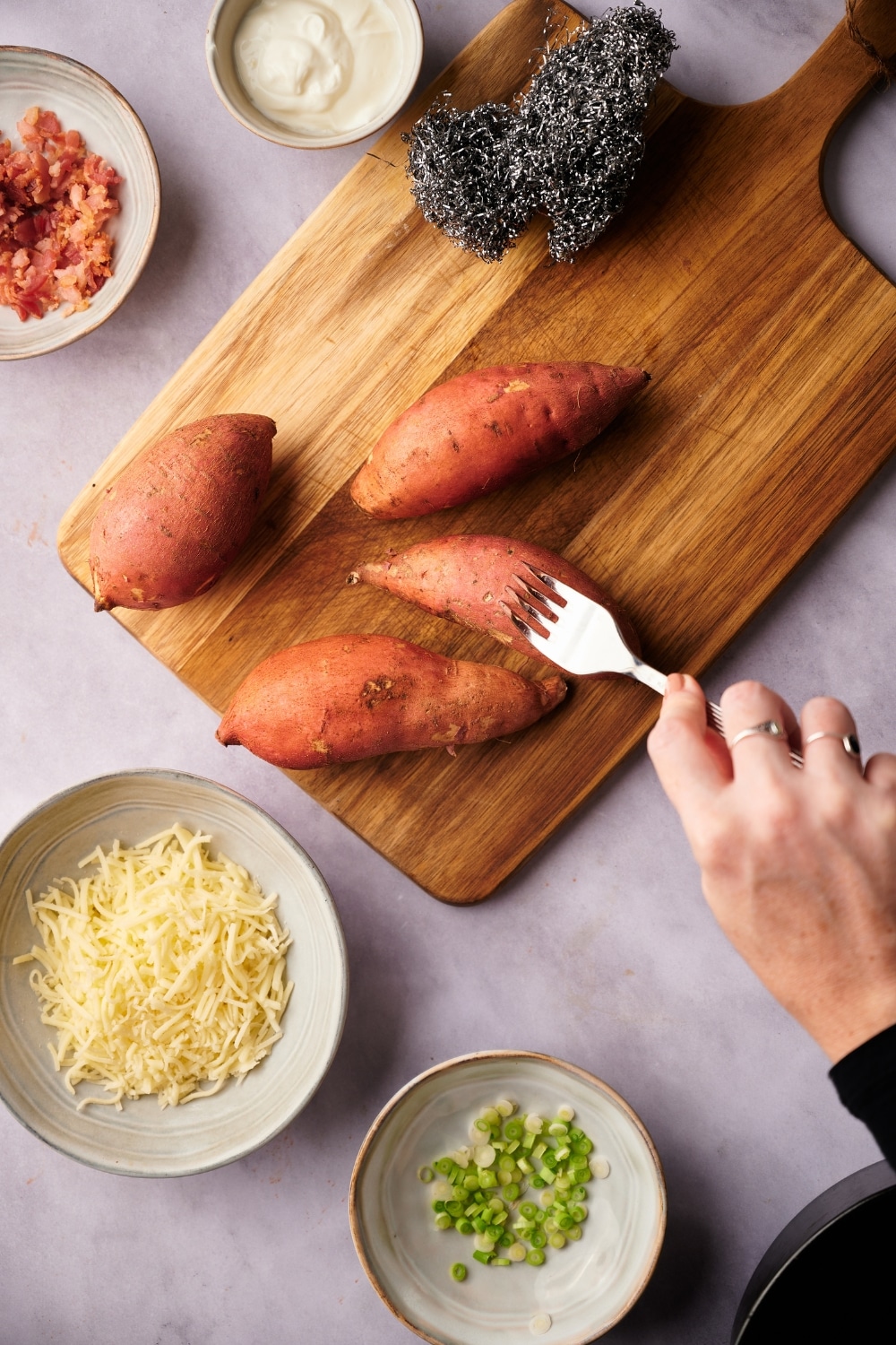 Four sweet potatoes on a wooden cutting board next to a steel wool scourer. A hand is piercing one of the sweet potatoes with a metal fork. Surrounding the cutting board are small ecru bowls of shredded mozzarella cheese, chopped green onions, bacon bits, and sour cream.