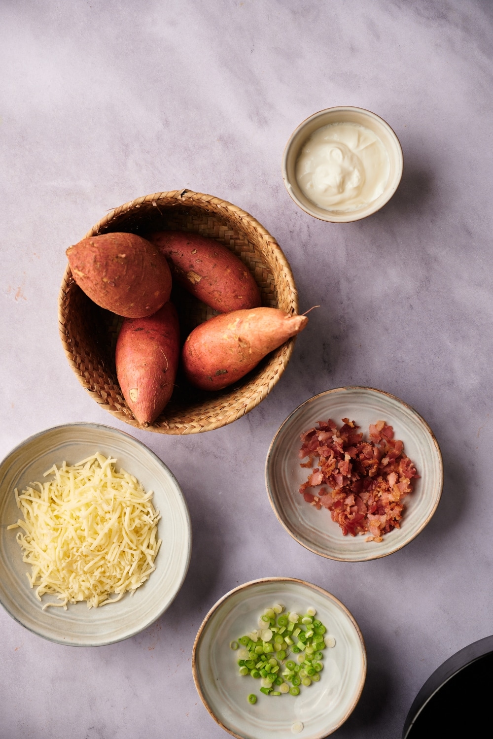 A woven rattan bowl with for sweet potatoes and small ecru bowls of shredded mozzarella cheese, chopped green onions, bacon bits, and sour cream.