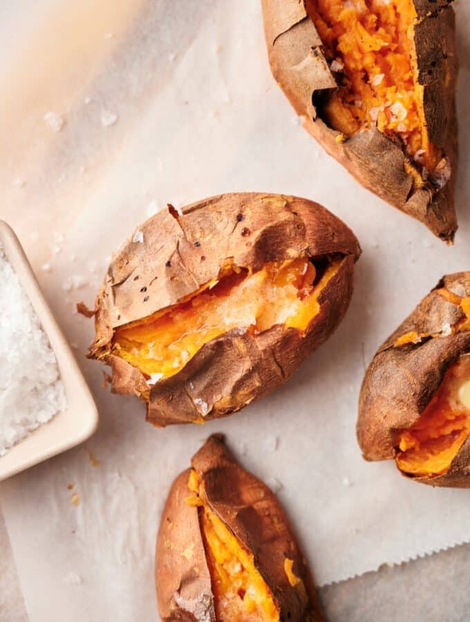 Close up of baked sweet potatoes cut open and arranged on a parchment paper lined wooden cutting board. Part of a square dipping bowl filled with salt is on the side.