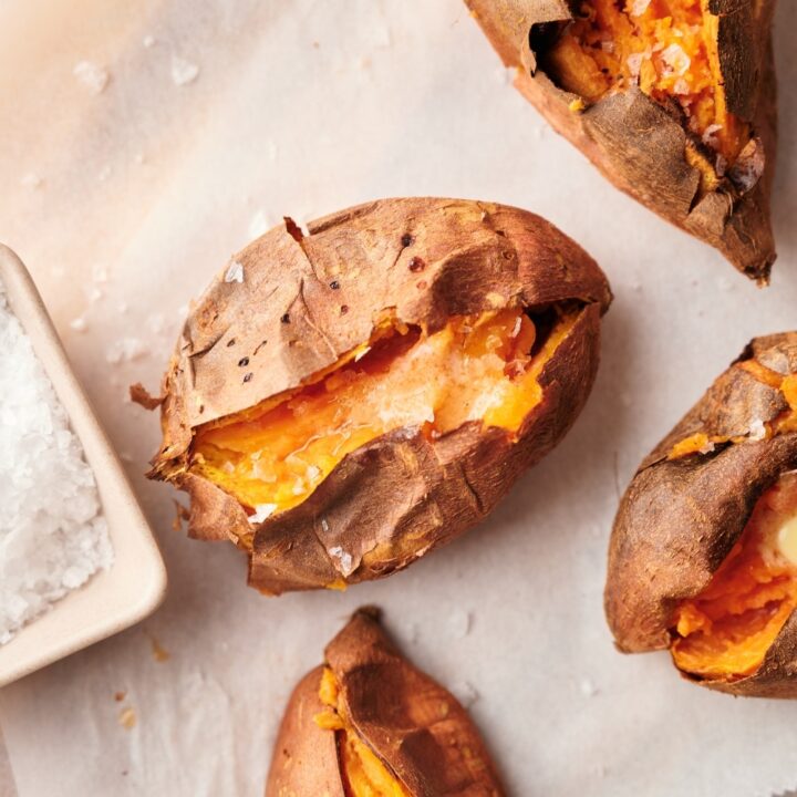 Close up of baked sweet potatoes cut open and arranged on a parchment paper lined wooden cutting board. Part of a square dipping bowl filled with salt is on the side.