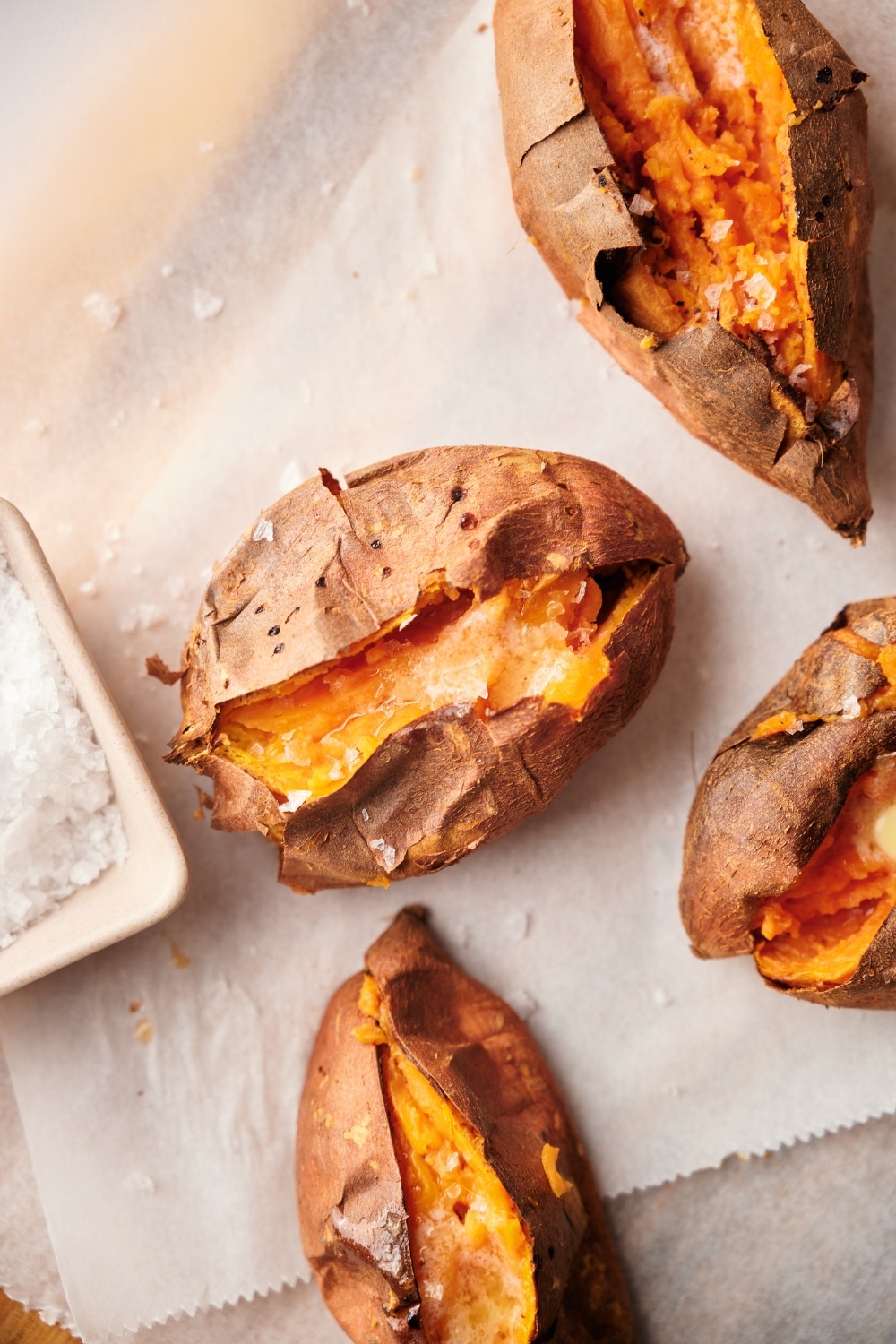 Close up of baked sweet potatoes cut open and arranged on a parchment paper lined wooden cutting board. Part of a square dipping bowl filled with salt is on the side.