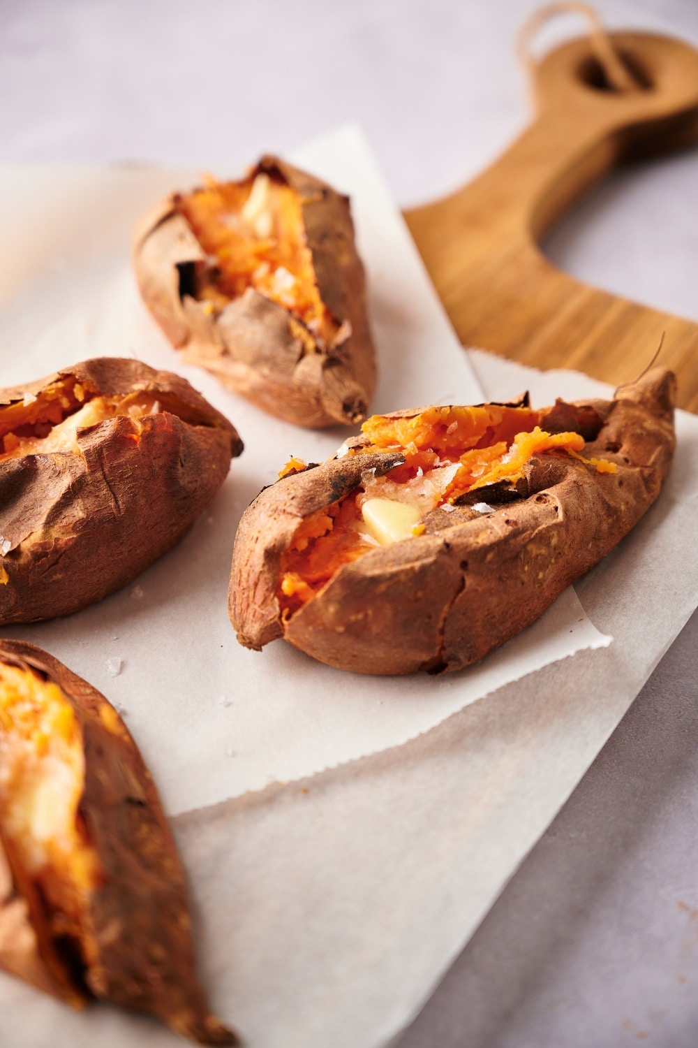 Four baked sweet potatoes cut open and arranged on a parchment paper lined wooden cutting board. A small piece of butter is melting in the center of each cut sweet potato.