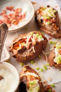 Close up of baked sweet potatoes garnished with shredded mozzarella cheese, bacon bits, sour cream, and chopped green onions. The sweet potatoes are served on parchmennt paper surrounded with small bowls of bacon bits, sour cream, and salt.