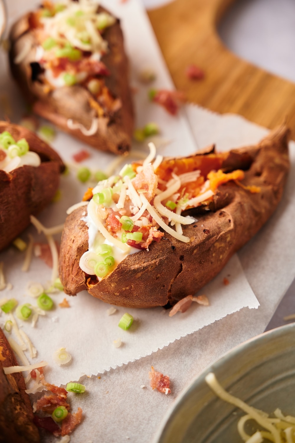 Close up of a baked sweet potato garnished with shredded mozzarella cheese, bacon bits, sour cream, and chopped green onions. The sweet potato is served with three more baked sweet potatoes on a parchment paper lined wooden cutting board.