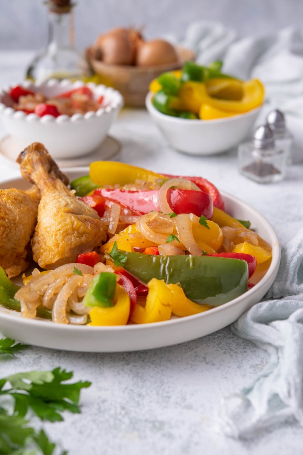 Sauteed bell peppers and onions garnished with parsley and served with fried chicken on a white plate. More sliced bell peppers and whole yellow onions can be seen in bowls in the back.