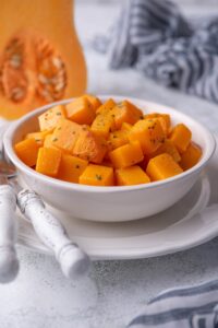 Sauteed butternut squash cubes in a white bowl. The bowl is on a white plate next to a metal fork and knife with white handles. In the back is a halved butternut squash and a tea towel.