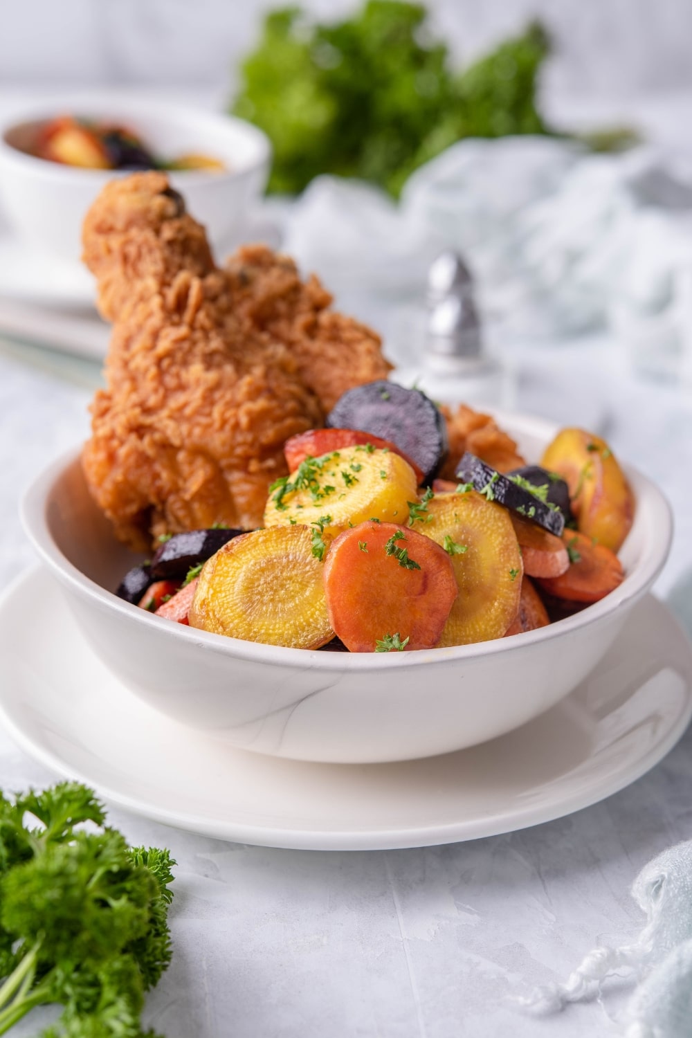 Sauteed carrots garnished with chopped parsley in a white bowl with breaded fried chicken. The bowl is on top of a white plate. Behind it is another bowl of sauteed carrots, a pair of salt and pepper shakers, and a bunch of fresh parsley.