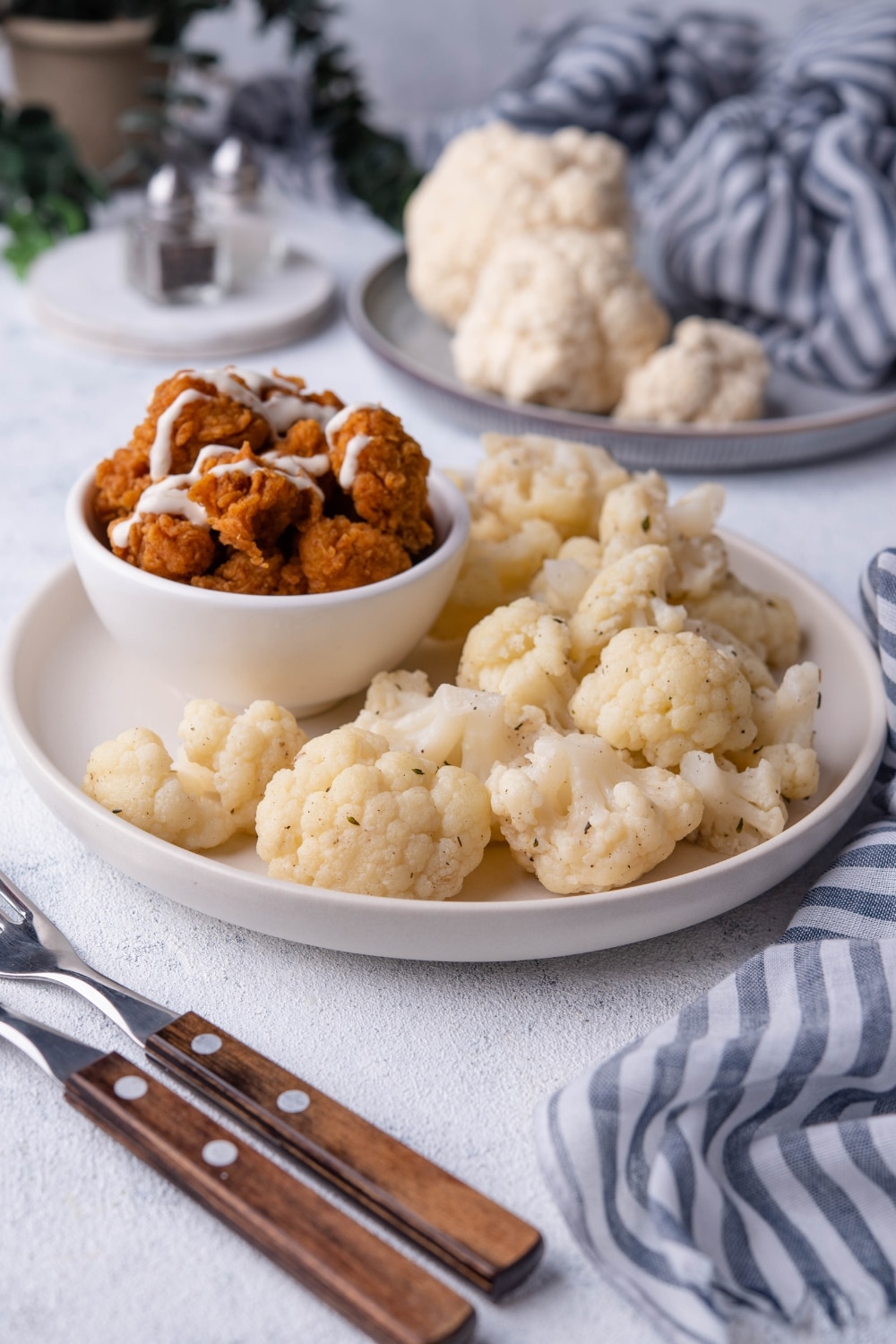 Sauteed cauliflower florets and a small bowl of fried chicken drizzled with white sauce on a white plate. Next to the plate are two forks with wooden handles and behind it is a plate of raw cauliflower and a pair of salt and pepper shakers.