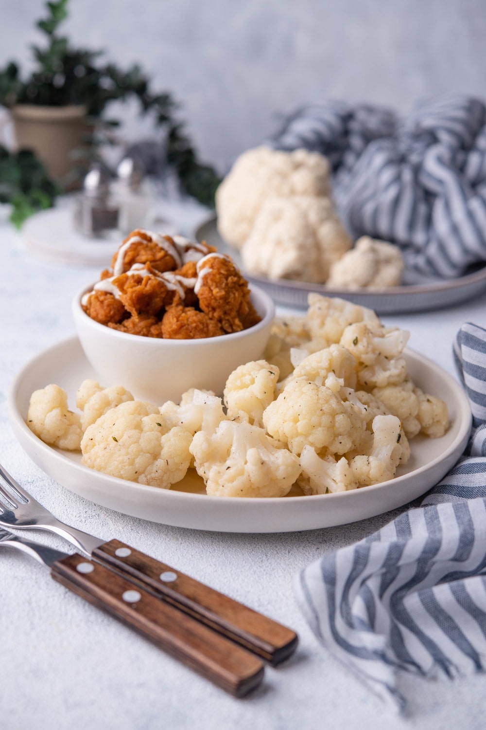 Sauteed cauliflower on a white plate with a small bowl of fried chicken drizzled with white sauce. Next to the plate are two forks with wooden handles and behind it is a plate of raw cauliflower, a draped tea towel, and a pair of salt and pepper shakers.